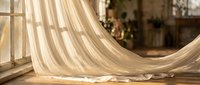 Flowing cream curtains with warm sunlight streaming through window creating soft shadows on wooden floor in cozy home interior for relaxation and comfort.