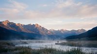 Misty mountain valley with serene river landscape at dawn, featuring layered peaks and morning fog over water for nature and travel projects.