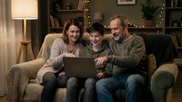 Happy family with teenage son using laptop together on cozy living room sofa in warm evening light. Parents and child bonding over technology at home.