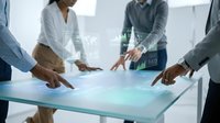 Diverse business team interacting with futuristic glowing touchscreen table displaying data charts and world map in modern office.