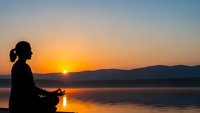 Woman silhouette meditating in lotus pose at sunrise by calm lake with mountains. Peaceful yoga practice, mindfulness and spiritual wellness concept.