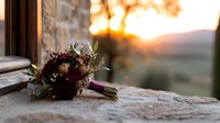 Wedding bouquet with dark burgundy dahlias and greenery resting on stone windowsill at sunset, romantic countryside view through window creates dreamy atmosphere.