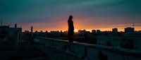 Young woman standing on urban rooftop at sunset, silhouetted against dramatic orange and teal sky with city skyline. Contemplative mood for lifestyle and inspirational content.