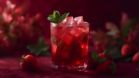 Refreshing strawberry cocktail with ice and mint garnish in glass on dark red background. Fresh berries scattered around for summer drink presentation.