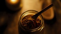 Dark cocoa powder in glass jar with spoon on moody background, warm lighting creates rich shadows and depth for food photography and culinary designs.