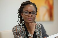 Professional African American businesswoman with glasses and dreadlocks in modern office setting, hand on chin in thoughtful pose during meeting.