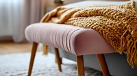 Cozy pink upholstered bench with wooden legs and chunky knit mustard throw blanket in modern interior setting against soft window light and carpet.