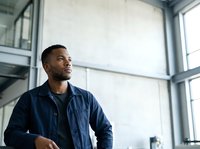 African American male professional in casual business attire gazing thoughtfully through large windows in modern office space, suitable for corporate, leadership, and workplace concepts.
