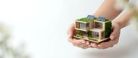 Hands holding miniature eco-friendly house model with green roof and solar panels representing sustainable architecture and environmental responsibility.