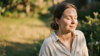 Middle-aged woman with dark hair in casual beige shirt standing outdoors in natural sunlight with blurred green garden background, eyes closed in peaceful moment, suitable for wellness and lifestyle content.