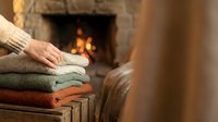 Person's hand resting on stack of folded knit sweaters near fireplace with warm flames glowing in background, cozy home interior setting.