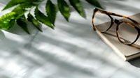 Stylish tortoiseshell eyeglasses resting on open book with green fern leaves and natural window light shadows creating elegant reading atmosphere.