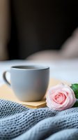 Cozy morning scene with gray ceramic cup of coffee, delicate pink rose on knitted blanket background, soft natural lighting creates romantic atmosphere.