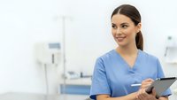 Young female healthcare professional in blue scrubs holding tablet in modern medical facility with equipment background for healthcare technology.
