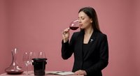 Professional female sommelier in black blazer tasting red wine at elegant table with decanter and glasses on pink background for wine education content.
