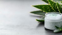 Fresh mint-colored face mask in glass jar with aloe vera leaves on gray concrete background, natural organic skincare and spa treatment concept.