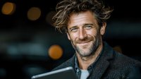 Mature Caucasian man with tousled gray hair and stubble smiling warmly at camera while wearing dark jacket, bokeh lights in background create atmospheric portrait.