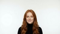 Young redhead woman with long wavy hair wearing black turtleneck sweater smiling at camera against white background for professional headshot photography.