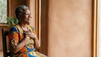 Mature African woman wearing traditional colorful patterned clothing, sitting indoors with warm natural light, expressing joy and contentment for lifestyle and cultural representation.
