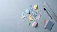 Colorful paper Easter bunny and egg cutouts with crafting supplies on gray background for spring holiday DIY projects and decorations.