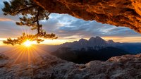 Dramatic mountain sunrise through natural rock arch frame, sunburst behind pine tree, dramatic orange cliffs and distant peaks against moody sky at dawn.
