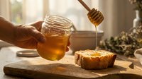 Unrecognizable person holding jar of golden honey with dripping dipper above honeycomb on wooden board, natural light, food styling.