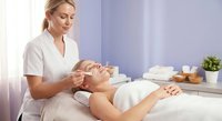 Professional blonde Caucasian female aesthetician performing facial treatment on relaxed client in modern spa wellness center with natural lighting.