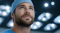 Young Caucasian male surgeon with blue eyes and beard wearing surgical cap in modern operating room with bright medical lights overhead.