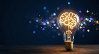 Glowing light bulb with brain-shaped filament on wooden surface against dark background with colorful bokeh lights representing innovation and creativity.