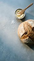 Freshly baked sourdough loaf with crusty exterior beside wooden bowl of creamy butter and scattered sea salt on blue-gray surface, ideal for bakery, food styling, and culinary content.