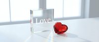 Crystal glass cube engraved with Love text beside a red gem heart on white surface, romantic Valentine concept with soft light.