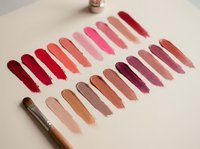 Colorful lipstick swatches arranged in rows showing various shades from red to pink to nude tones with makeup brush on neutral background.
