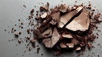 Crushed brown eyeshadow or bronzer powder with scattered fragments on neutral background, showing makeup texture and pigmentation.