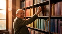 Elderly Caucasian man with glasses reaching for book on library shelf surrounded by extensive collection of volumes in warm natural lighting for education concepts.
