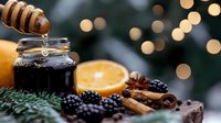 Honey dripping into jar with mulled wine ingredients including orange, cinnamon sticks, star anise and blackberries against festive bokeh lights background.