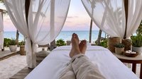 Relaxed woman lying on bed with feet up under white canopy curtains overlooking serene ocean beach at sunset, luxury resort spa wellness retreat destination.
