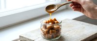 Hand drizzling golden caramel sauce over roasted nuts in glass bowl on wooden cutting board near bright window for gourmet dessert preparation.