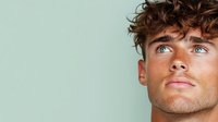 Young Caucasian male model with curly brown hair and blue eyes looking up against mint green background, close-up portrait showing defined facial features and natural lighting.