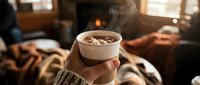 Hands holding a steaming cup of hot cocoa with marshmallows in a cozy cabin interior with fireplace in background.