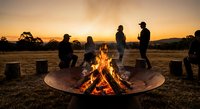 Group of friends gathering around campfire at sunset in rural field with warm golden sky and silhouetted figures enjoying outdoor evening together.