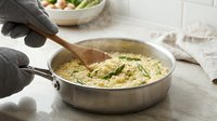 Person in black gloves stirring creamy pasta sauce with wooden spoon in stainless steel pan, fresh herbs garnish, kitchen preparation scene for cooking content.