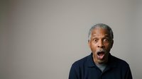 Senior man with gray hair and dark skin, wearing navy polo shirt, expressing surprise or shock with open mouth against neutral gray background, suitable for business, healthcare, and lifestyle editorial use.