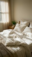 Cozy minimalist bedroom with unmade white linen bedding, natural morning light through sheer curtains, wooden nightstand with coffee cup and plant.