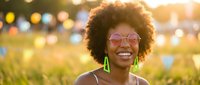 Young African American woman with afro hair smiling at outdoor music festival, wearing pink sunglasses and green earrings in golden sunlight.