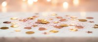 Golden confetti scattered on white surface with warm bokeh lights creating festive celebration atmosphere for party decorations and events.