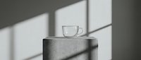 Clear glass cup with handle placed on concrete cylinder with natural sunlight casting geometric shadows on neutral background.