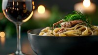 Creamy pasta with mushrooms and fresh dill in elegant black bowl, accompanied by red wine glass with warm bokeh lights creating cozy dining atmosphere.
