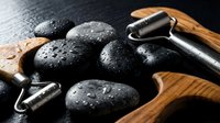 Black spa stones with water droplets and stainless steel facial roller on wooden tray for relaxation massage therapy and skincare treatment.