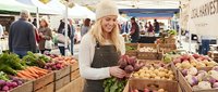 Young blonde woman farmer in beige knit hat and apron selling fresh organic vegetables at bustling outdoor farmers market with wooden crates full of produce.