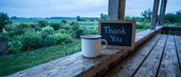 Rustic wooden porch with enamel mug and chalkboard sign reading Thank You, overlooking lush green garden at dusk, gratitude concept.